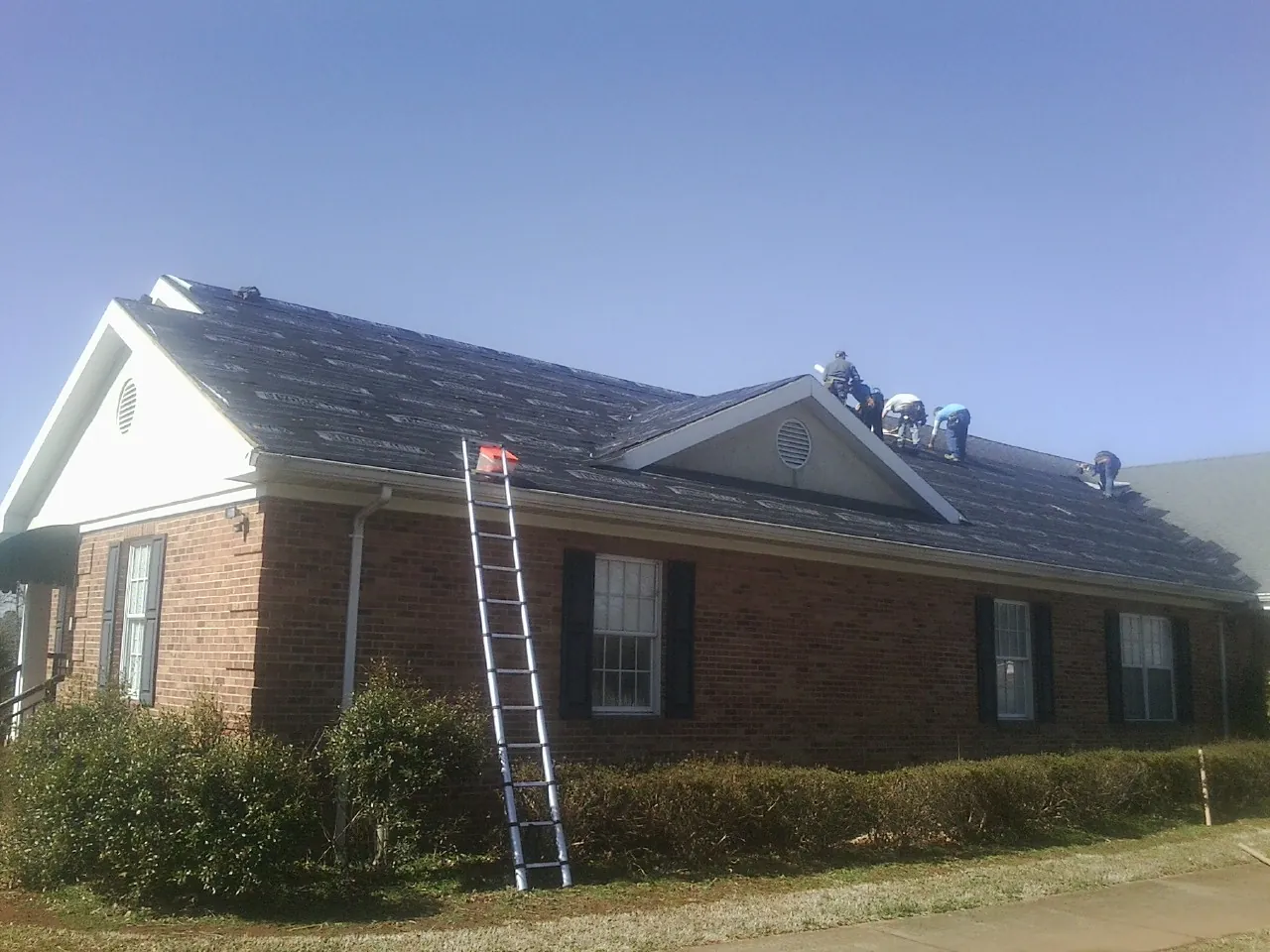 Crew tearing shingles off a brick ranch roof with a ladder set — Metal Roof Repair work by our crew in Lamoure