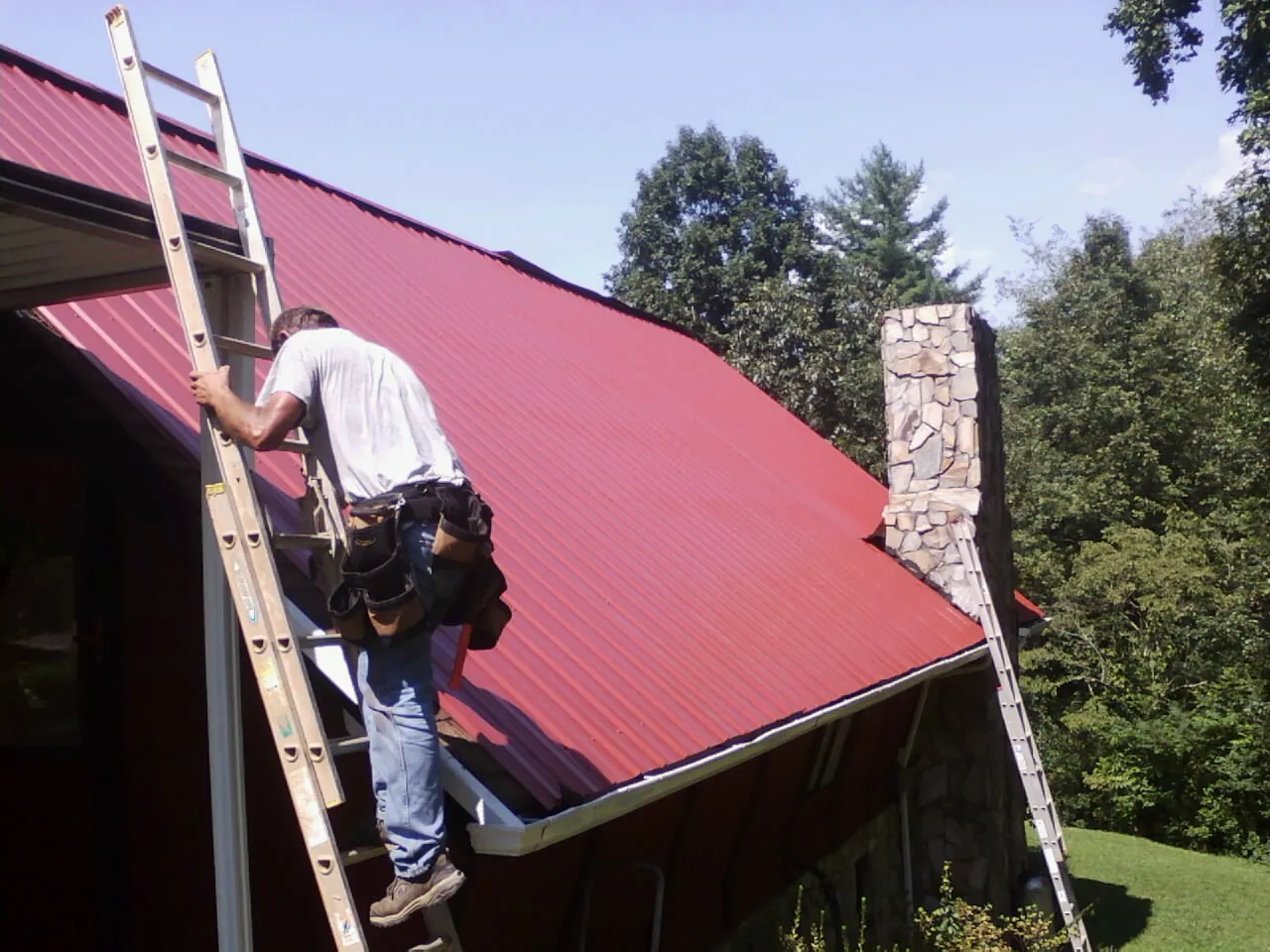 Roofer on a ladder beside a red metal roof and stone chimney — Soffit & Fascia Repair by Roofing Brothers in Lamoure